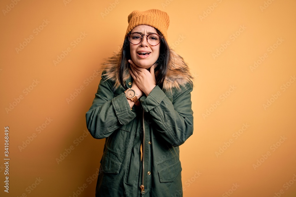 Young brunette woman wearing glasses and winter coat with hat over yellow isolated background shouting and suffocate because painful strangle. Health problem. Asphyxiate and suicide concept.