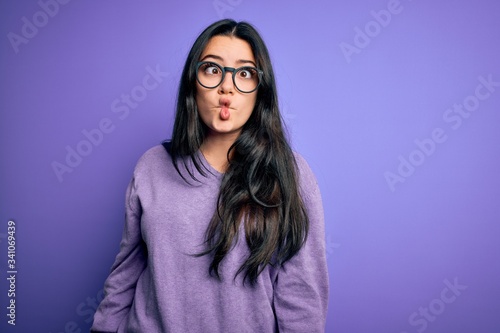 Young brunette woman wearing glasses over purple isolated background making fish face with lips, crazy and comical gesture. Funny expression.