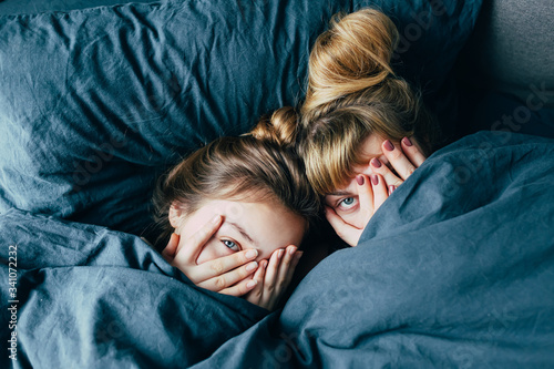 Portrait of light hair long hair mother and daughter under the duvet together in soft morning light on blue linen bed. Concept of happy family living, relaxation, comfort, fun. Play hide and seek