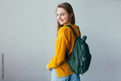 teenage girl looking at camera and smiles. Beautiful student girl standing in yellow sweater and blue jeans and green backpack dressed on shoulders. Trendy casual outfit for teen woman. hands in pock
