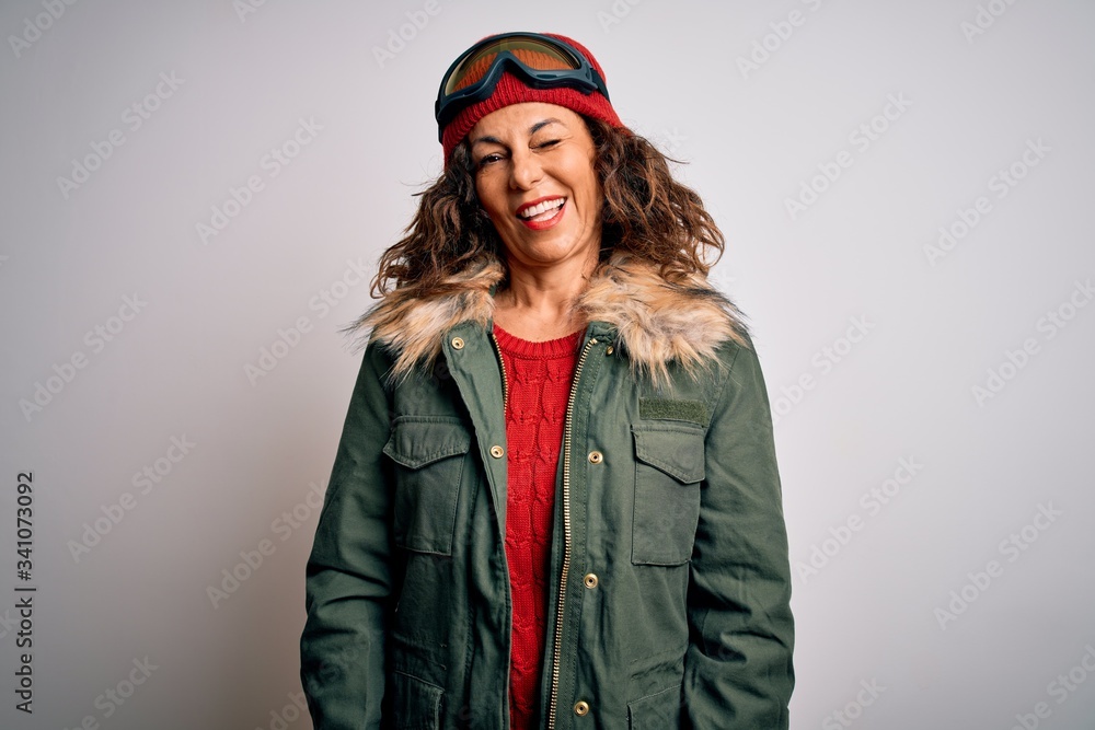 Middle age skier woman wearing snow sportswear and ski goggles over white background winking looking at the camera with sexy expression, cheerful and happy face.
