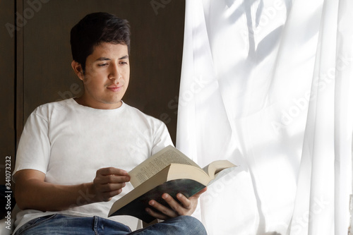 Young mexican man reading next to the window with soft ligth