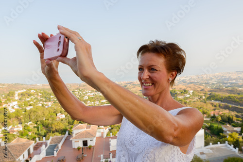 Happy mature tourist woman taking picture with phone of the beautiful scenery of Mijas village in Spain