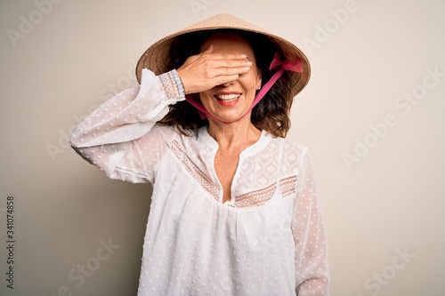 Middle age brunette woman wearing asian traditional conical hat over white background smiling and laughing with hand on face covering eyes for surprise. Blind concept.