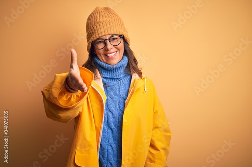 Middle age woman wearing yellow raincoat and winter hat over isolated background smiling friendly offering handshake as greeting and welcoming. Successful business. © Krakenimages.com