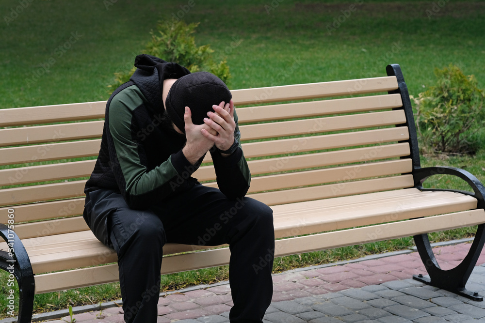 A guy in black pants, a sports jacket and a black hat sits on a bench. A man sits on a wooden bench bowing his head and holding it with his hands. The guy on the bench in frustrated feelings, despair