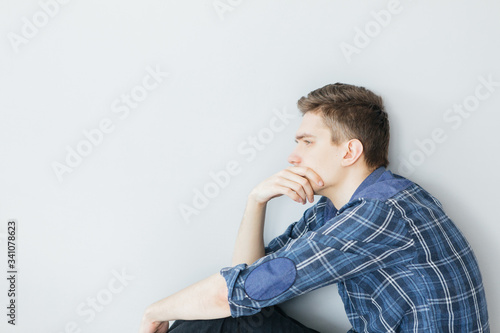 A young man is sitting near a light gray wall. Pensive young man in a blue shirt © alexander132