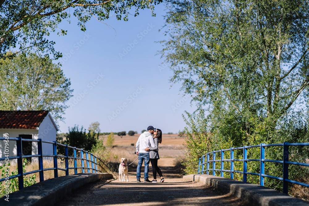 A kissing couple with their dog standing on a bridge in a park A kissing couple with their dog standing on a bridge in a park