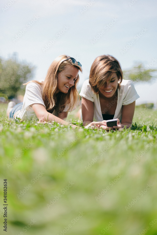 Two smiling female friends lying on a meadow sharing cell phone Two smiling female friends lying on a meadow sharing cell phone