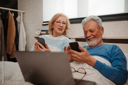 Portrait of smiling couple lying in bed and browsing social networks together at their smartphones.