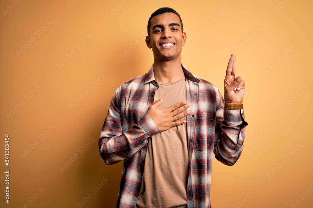 Young handsome african american man wearing casual shirt standing over yellow background smiling swearing with hand on chest and fingers up, making a loyalty promise oath Young handsome african american man wearing casual shirt standing over yellow background smiling swearing with hand on chest and fingers up, making a loyalty promise oath