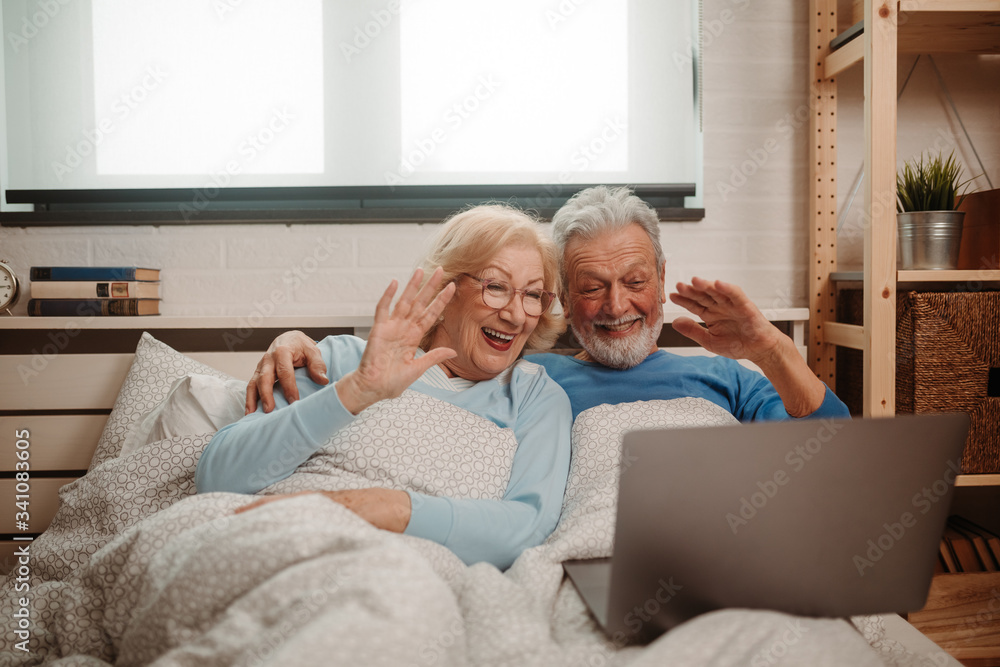 Portrait of lovely couple lying in bed and having Skype video call. Elderly couple waving to their grandchildren during video call. Portrait of lovely couple lying in bed and having Skype video call. Elderly couple waving to their grandchildren during video call.
