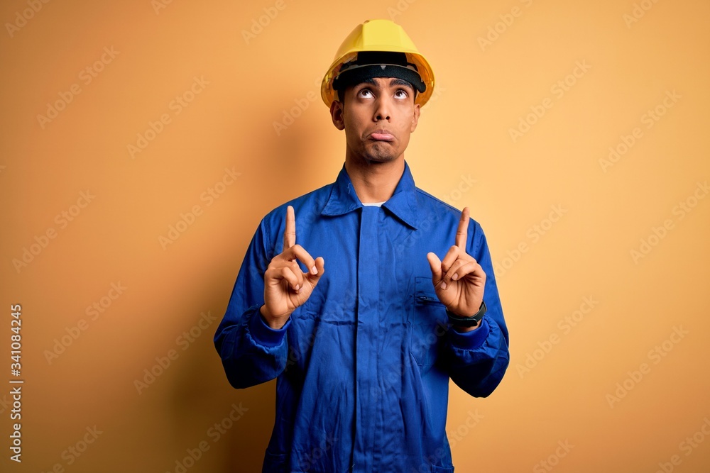 Young handsome african american worker man wearing blue uniform and security helmet Pointing up looking sad and upset, indicating direction with fingers, unhappy and depressed.