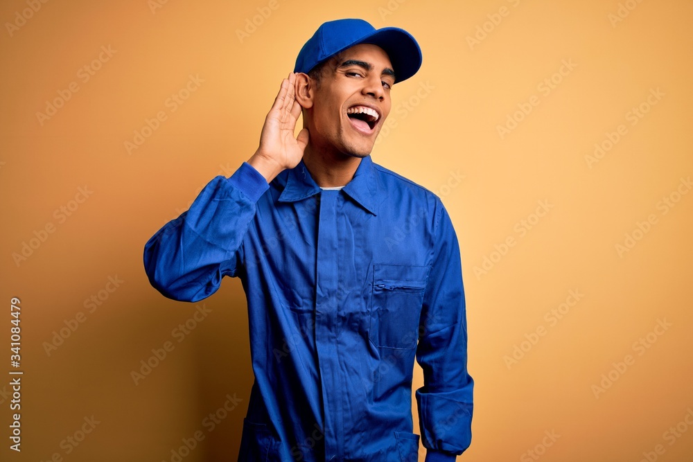 Young african american mechanic man wearing blue uniform and cap over yellow background smiling with hand over ear listening an hearing to rumor or gossip. Deafness concept.