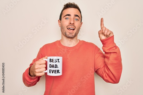 Young handsome man with blue eyes drinking cup of coffe with best dad ever message surprised with an idea or question pointing finger with happy face, number one