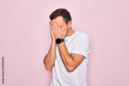 Handsome man with blue eyes wearing casual white t-shirt standing over pink background with sad expression covering face with hands while crying. Depression concept.