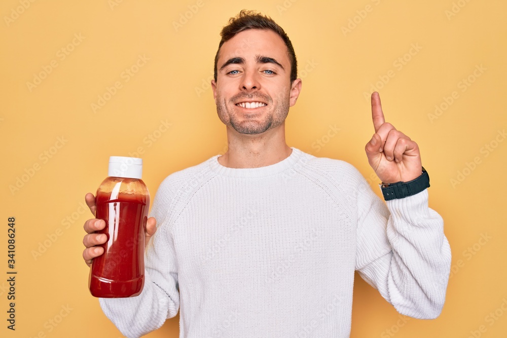 Young handsome man holding bottle of ketchup sauce condiment over yellow background surprised with an idea or question pointing finger with happy face, number one Young handsome man holding bottle of ketchup sauce condiment over yellow background surprised with an idea or question pointing finger with happy face, number one