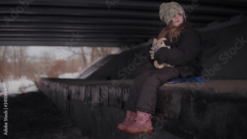 A helpless scared little homeless girl in a holey knitted multi-colored hat sits in the winter under the bridge. The child hugs a single beige soft toy