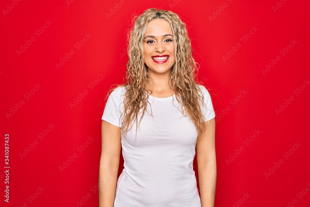 Young beautiful blonde woman wearing casual t-shirt standing over isolated red background with a happy and cool smile on face. Lucky person. Young beautiful blonde woman wearing casual t-shirt standing over isolated red background with a happy and cool smile on face. Lucky person.