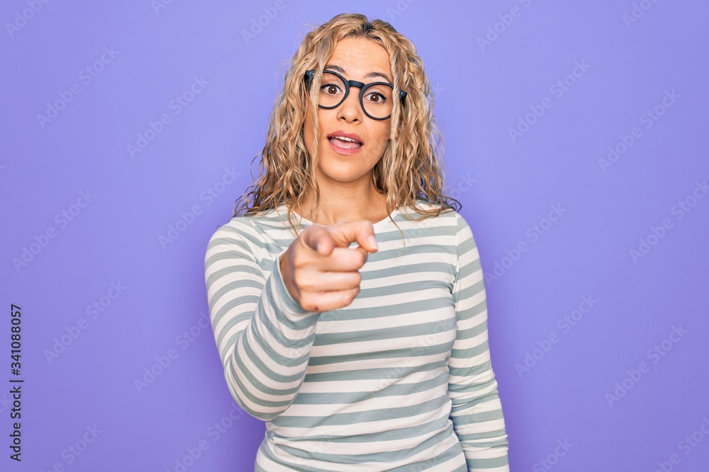 Beautiful blonde woman wearing casual striped t-shirt and glasses over purple background pointing displeased and frustrated to the camera, angry and furious with you Beautiful blonde woman wearing casual striped t-shirt and glasses over purple background pointing displeased and frustrated to the camera, angry and furious with you