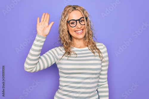 Wallpaper Mural Beautiful blonde woman wearing casual striped t-shirt and glasses over purple background Waiving saying hello happy and smiling, friendly welcome gesture Torontodigital.ca