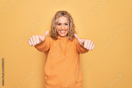 Young beautiful blonde sporty woman wearing casual sweatshirt over yellow background approving doing positive gesture with hand, thumbs up smiling and happy for success. Winner gesture.