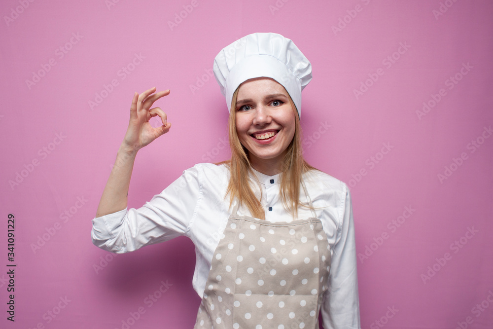 beautiful cheerful girl cook in uniform and apron on a colored pink background shows okay, portrait of a housewife woman in kitchen clothes beautiful cheerful girl cook in uniform and apron on a colored pink background shows okay, portrait of a housewife woman in kitchen clothes