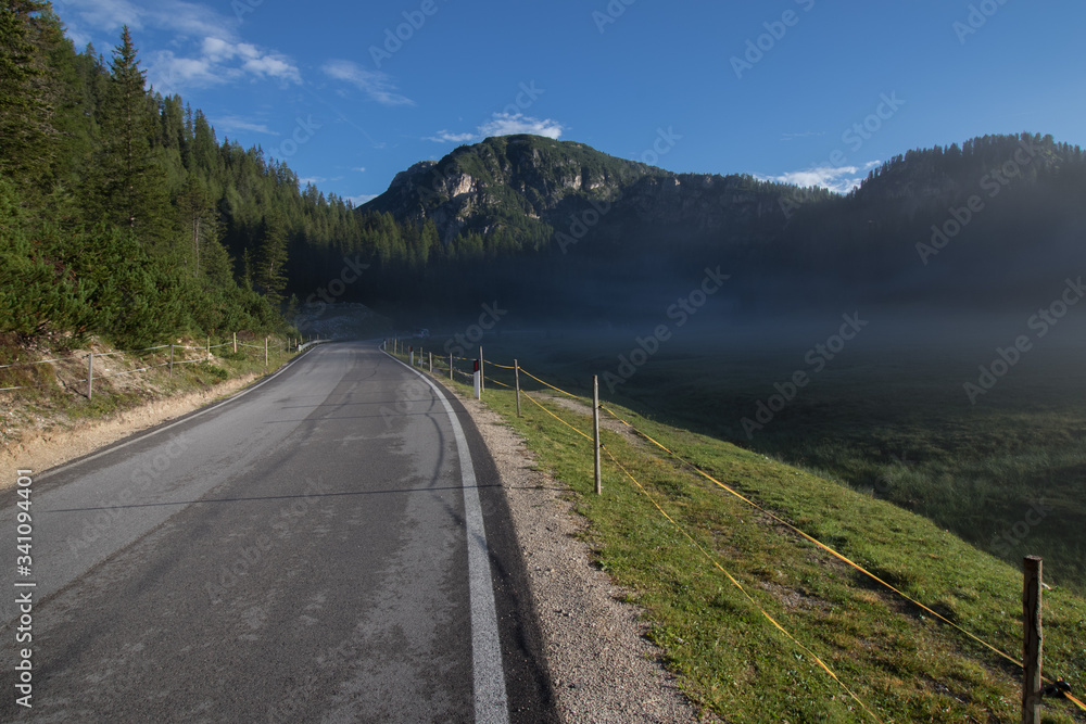 Obraz premium A small, light morning fog rising above the meadow by the asphalt road. Dolomite, Italy.