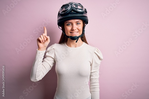 Young beautiful motorcyclist woman with blue eyes wearing moto helmet over pink background with a big smile on face, pointing with hand finger to the side looking at the camera.