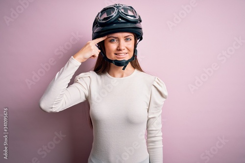 Young beautiful motorcyclist woman with blue eyes wearing moto helmet over pink background Smiling pointing to head with one finger, great idea or thought, good memory © Krakenimages.com