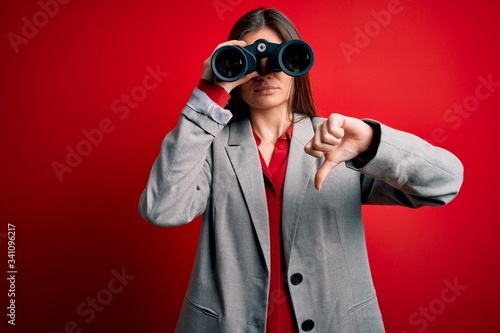 Young beautiful woman with blue eyes using binoculars over isolated red background with angry face, negative sign showing dislike with thumbs down, rejection concept