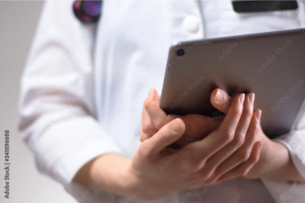 Tablet in the hands of healthcare professional up close. White coat, stethoscope, and badge visible in background. Hands of nurse practitioner or PA using technology in medicine for patients Tablet in the hands of healthcare professional up close. White coat, stethoscope, and badge visible in background. Hands of nurse practitioner or PA using technology in medicine for patients