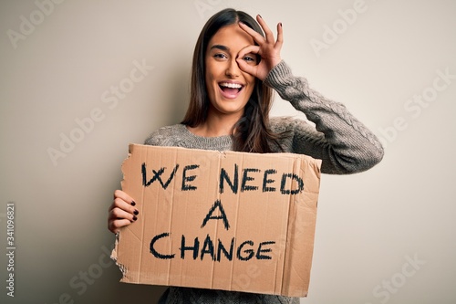 Young beautiful brunette activist woman protesting for a change holding banner with happy face smiling doing ok sign with hand on eye looking through fingers © Krakenimages.com