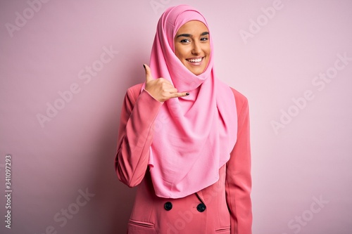 Young beautiful brunette businesswoman wearing pink muslim hijab and business jacket smiling doing phone gesture with hand and fingers like talking on the telephone. Communicating concepts. © Krakenimages.com