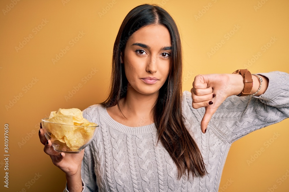 Young beautiful brunette woman holding bowl with snack potato chips over yellow background with angry face, negative sign showing dislike with thumbs down, rejection concept Young beautiful brunette woman holding bowl with snack potato chips over yellow background with angry face, negative sign showing dislike with thumbs down, rejection concept