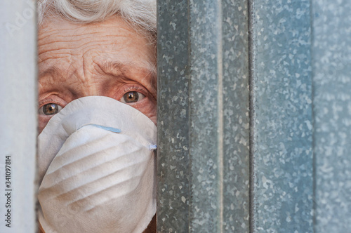 Принти на полотні A frightened grandmother in a mask peeps out of the gap in the fence through the gate of her house