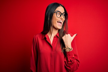 Young beautiful brunette woman wearing casual shirt and glasses over red background smiling with happy face looking and pointing to the side with thumb up.