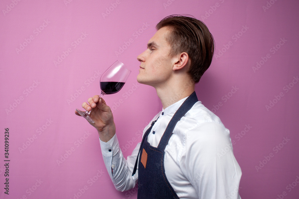 young sommelier guy checks the smell of red wine on a colored background, an expert on wine in a white shirt and apron holds a glass, the waiter tastes the wine young sommelier guy checks the smell of red wine on a colored background, an expert on wine in a white shirt and apron holds a glass, the waiter tastes the wine