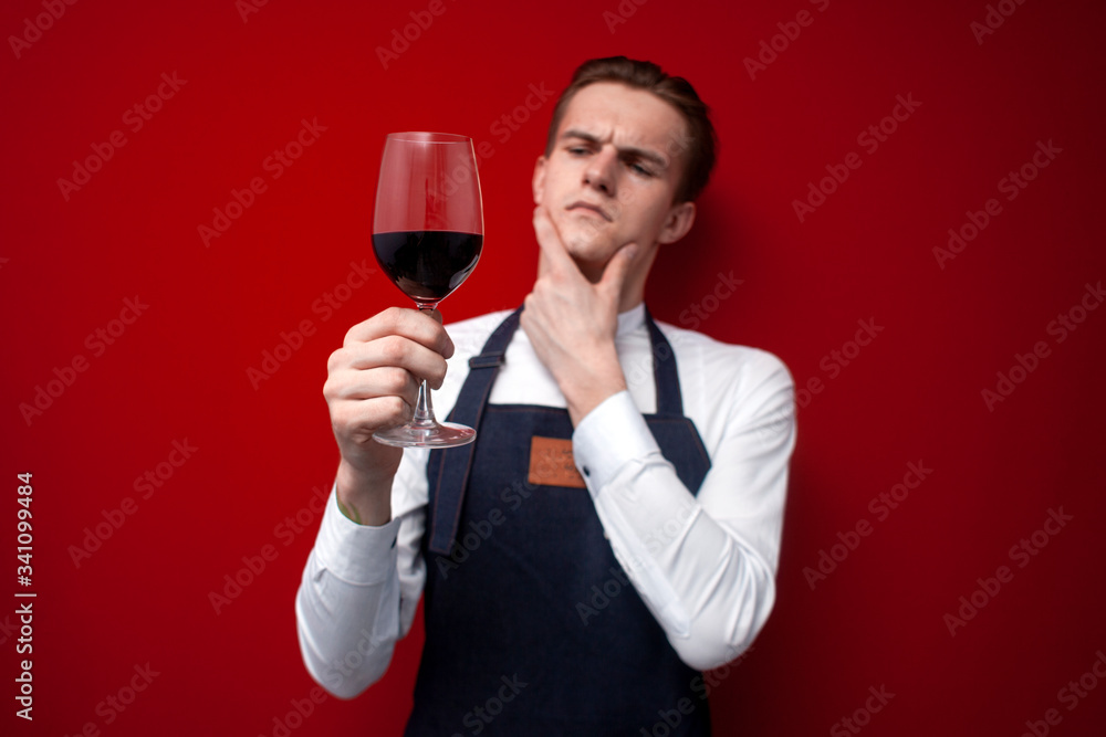 professional thoughtful sommelier holds a glass of red wine and thinks against a red background, the guy the waiter looks at the wine professional thoughtful sommelier holds a glass of red wine and thinks against a red background, the guy the waiter looks at the wine