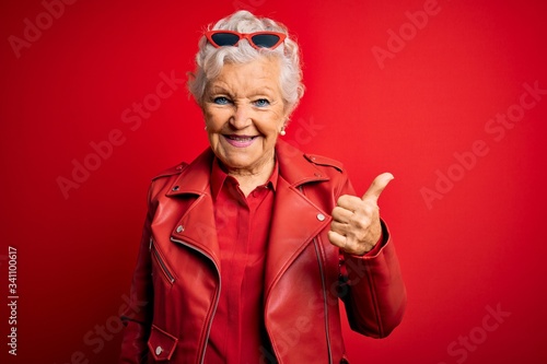 Senior beautiful grey-haired woman wearing casual red jacket and sunglasses doing happy thumbs up gesture with hand. Approving expression looking at the camera showing success.