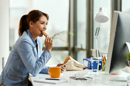 Fototapeta Smiling businesswoman eating a cookie while using mobile phone in the office