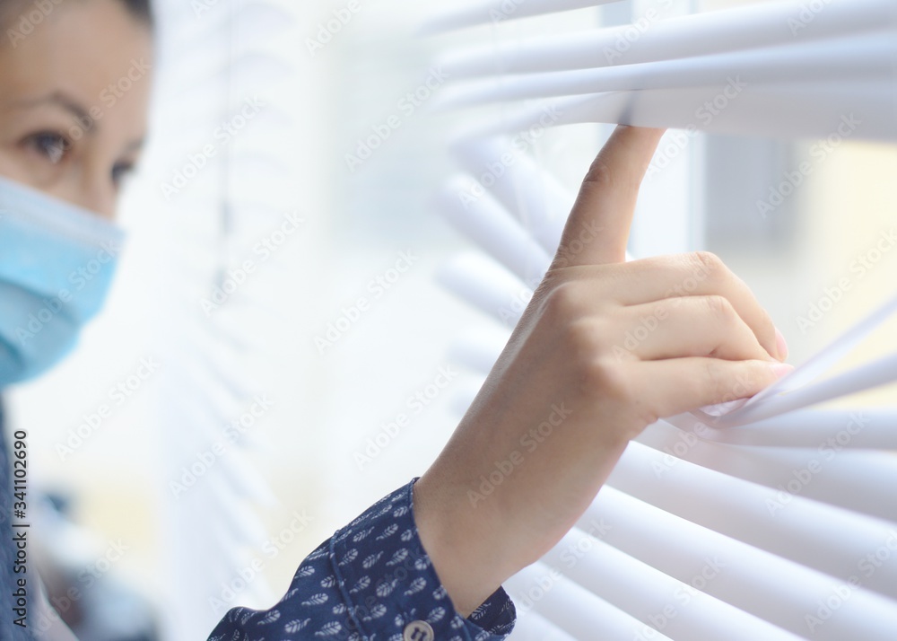 Mujer con tapabocas o mascara facial en cuarentena con su mano en la persiana o cortina mirando por una ventana Mujer con tapabocas o mascara facial en cuarentena con su mano en la persiana o cortina mirando por una ventana
