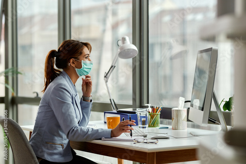 Pensive businesswoman with face mask reading an e-mail on desktop PC in the office.