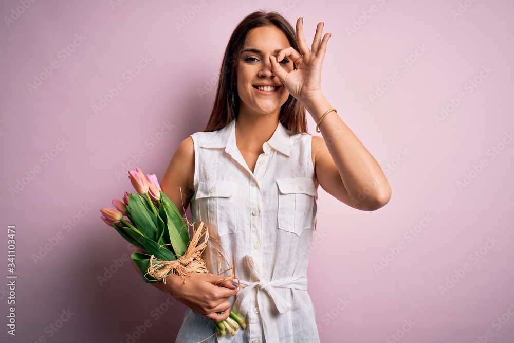 Young beautiful brunette woman holding bouquet of tulips flowers over pink background doing ok gesture with hand smiling, eye looking through fingers with happy face. Young beautiful brunette woman holding bouquet of tulips flowers over pink background doing ok gesture with hand smiling, eye looking through fingers with happy face.