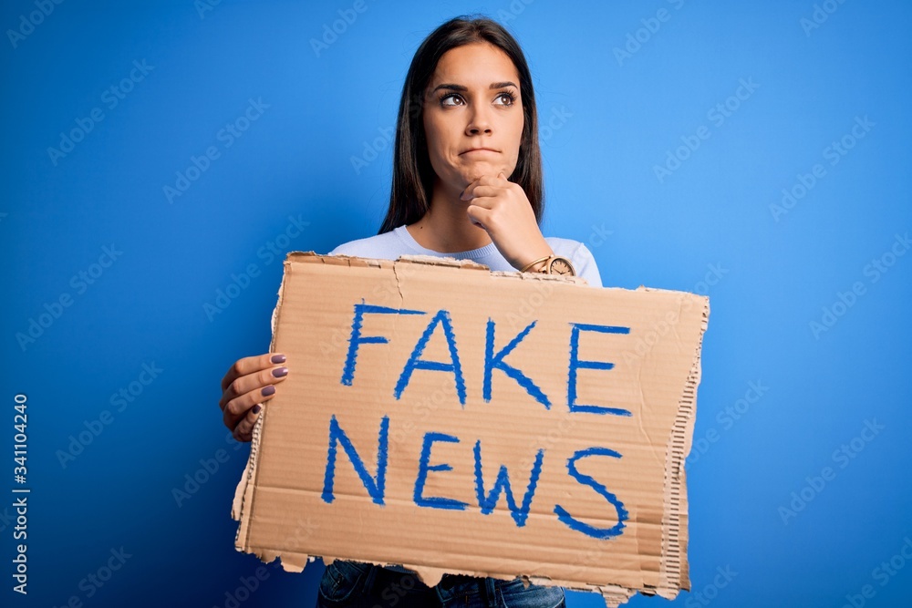 Young beautiful brunette woman holding banner with fake news message serious face thinking about question, very confused idea Young beautiful brunette woman holding banner with fake news message serious face thinking about question, very confused idea