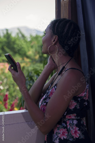 beautiful tanned young girl smiling and chatting with sell phone inside a house leaning in a window with view of outside