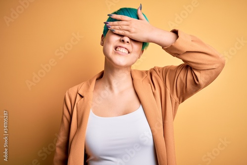 Young beautiful woman with blue fashion hair wearing casual jacket over yellow background smiling and laughing with hand on face covering eyes for surprise. Blind concept.