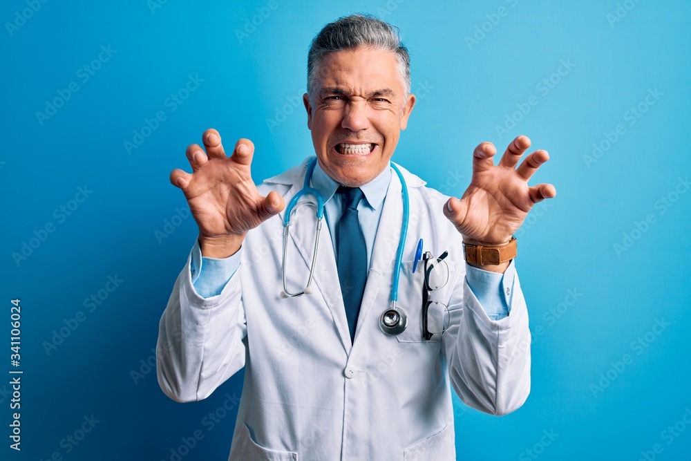 Middle age handsome grey-haired doctor man wearing coat and blue stethoscope smiling funny doing claw gesture as cat, aggressive and sexy expression Middle age handsome grey-haired doctor man wearing coat and blue stethoscope smiling funny doing claw gesture as cat, aggressive and sexy expression