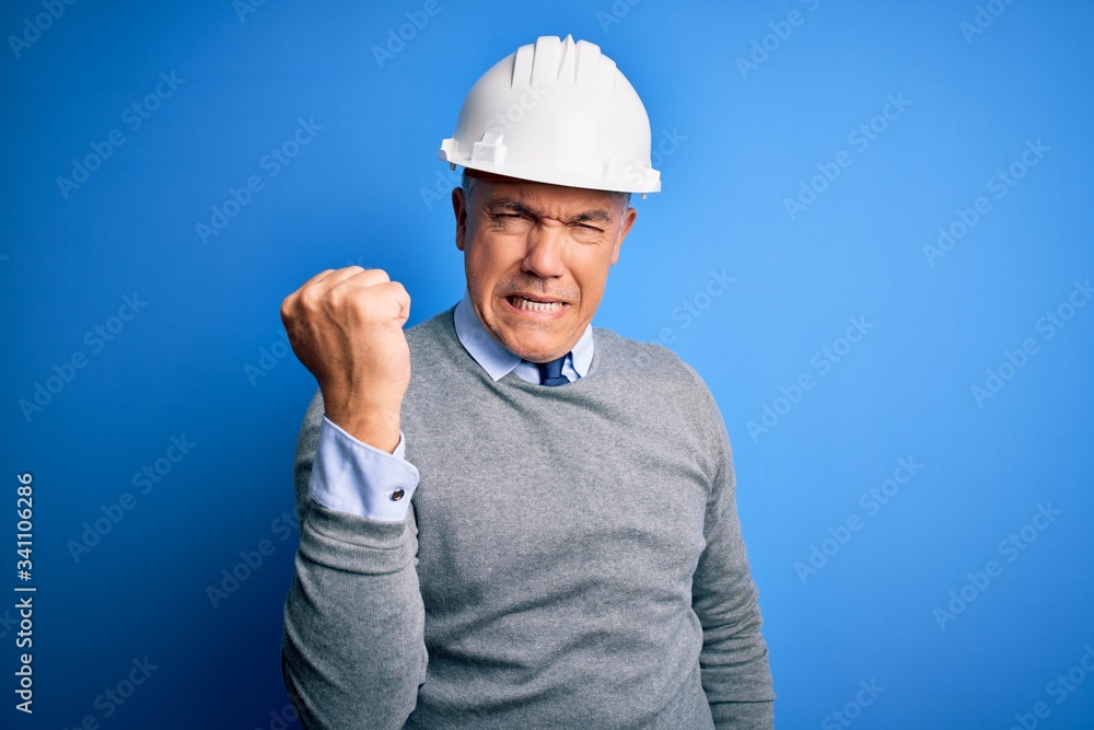 Middle age handsome grey-haired engineer man wearing safety helmet over blue background angry and mad raising fist frustrated and furious while shouting with anger. Rage and aggressive concept. Middle age handsome grey-haired engineer man wearing safety helmet over blue background angry and mad raising fist frustrated and furious while shouting with anger. Rage and aggressive concept.