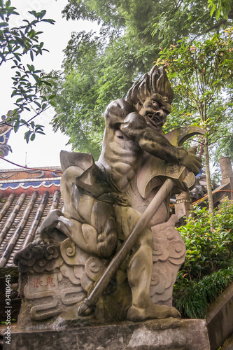 Fengdu, China - May 8, 2010: Ghost City, historic sanctuary. Closeup of brown stone hatchet holding man-like angry monster statue on wall under green foliage and silver sky.